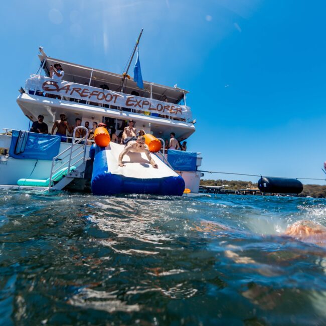 A large yacht named "Barefoot Explorer," filled with people, is anchored in the water on a sunny day. The yacht features an inflatable slide leading into the water. Other boats are visible, and the shore with trees can be seen in the background—a perfect scene for Boat Parties Sydney The Yacht Social Club.