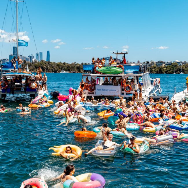 A vibrant, sunny day on the water with numerous people floating on colorful inflatables. A crowded boat labeled "Rum Runner Cruises" is in the background. The scene is lively with people swimming and enjoying themselves, and the city skyline visible in the distance hints at a perfect Sydney Harbour Boat Hire experience.