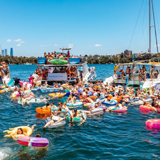 A large group of people enjoy a sunny day on the water, floating on colorful inflatables near several anchored boats. The scene is lively with people swimming, socializing, and basking in the sun against a backdrop of a city skyline and boats, reminiscent of Boat Parties Sydney by The Yacht Social Club.