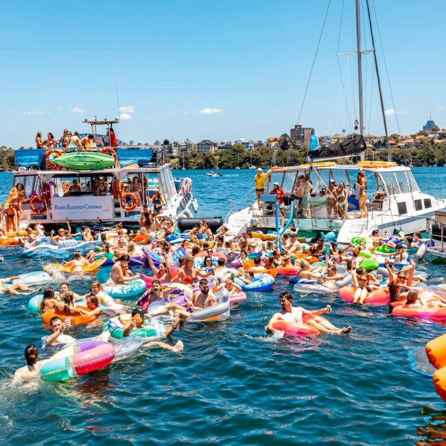 A vibrant scene of a yacht party on a sunny day, with numerous people floating on colorful inflatable rafts and tubes in the water. Several boats are anchored nearby as part of The Yacht Social Club Sydney Boat Hire, filled with partygoers enjoying the event. The skyline of a city is visible in the background.