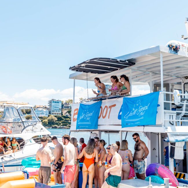 A lively group of people, mostly in swimwear, enjoying a sunny day on a yacht. They are gathered on the deck, with some standing and some sitting. The yacht features banners for "The Yacht Social Club Sydney Boat Hire" and is surrounded by other boats and inflatables.