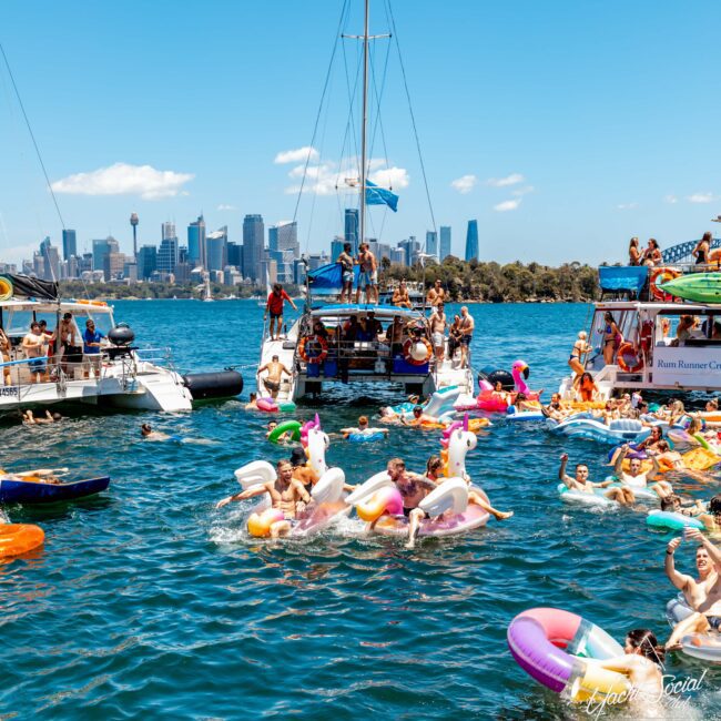 People are enjoying a sunny day on the water, floating on various colorful inflatables near several boats from The Yacht Social Club Event Boat Charters. The city skyline is visible in the background, and the atmosphere is lively with music and laughter. The scene includes unicorn and donut-shaped inflatables.