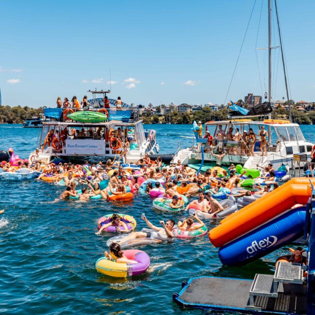 A large group of people enjoying a sunny day on boats and inflatable rafts in a body of water. Many individuals are in colorful floaties, with some dancing and socializing. Several boats from The Yacht Social Club Sydney Boat Hire are anchored nearby. The city skyline is visible in the background.