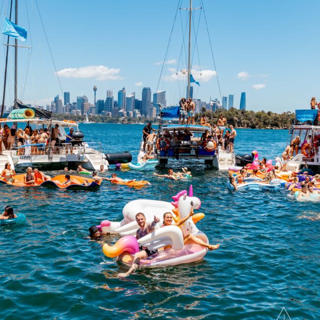 A group of people enjoys a sunny day on the water, with several floating toys and yachts in the background. Two individuals float on an inflatable unicorn. The city skyline is visible in the distance, and a "Yacht Social Club" logo is in the bottom right corner, promoting Boat Parties Sydney by The Yacht Social Club.