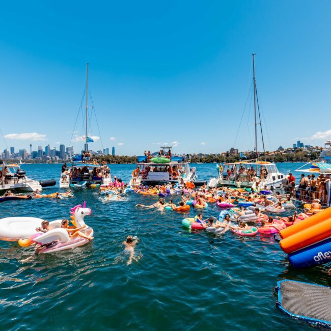 A lively scene on a sunny day features a large group of people in a marina. They are on various inflatable rafts and boats, enjoying the water. Several yachts and boats from The Yacht Social Club Sydney Boat Hire are anchored nearby, with the city skyline visible in the background.