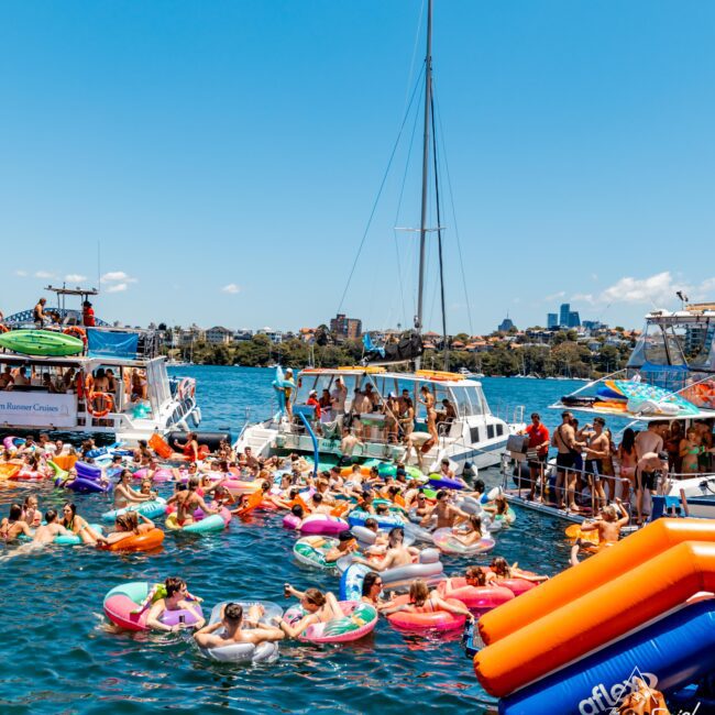 People enjoying a sunny day at The Yacht Social Club, floating in the water with colorful inflatable rings. Luxury yachts are docked nearby, and some people are seen standing and socializing on the boats. The backdrop includes a clear blue sky and distant cityscape.