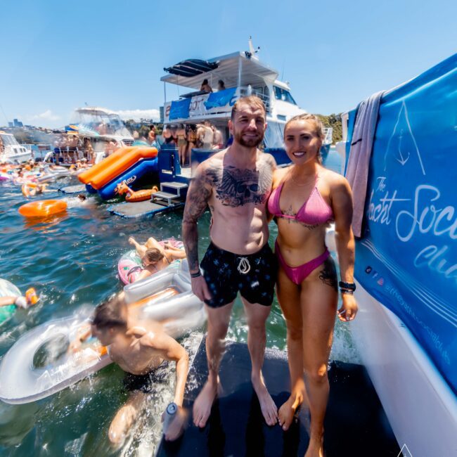 A man and woman, both in swimwear, pose on a floating platform beside a yacht on a sunny day. Other people enjoy the water and inflatable rings nearby. A banner on the yacht reads "The Yacht Social Club." The atmosphere is lively and festive, perfect for Boat Rental and Parties Sydney The Yacht Social Club offers.