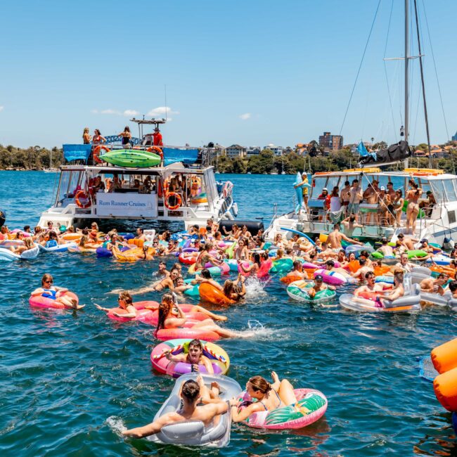 A vibrant scene of people in colorful inflatables enjoying a sunny day in the water near anchored boats. The clear blue sky and the distant shoreline with buildings add to the lively atmosphere, enhanced by Sydney Boat Hire from The Yacht Social Club. Some participants relax while others engage in playful activities.