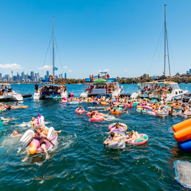 A lively boat party on a sunny day, with people enjoying themselves in the water on colorful inflatables. Multiple boats are anchored nearby, and a city skyline is visible in the distance. The scene is festive with a clear blue sky overhead, courtesy of The Yacht Social Club Sydney Boat Hire.