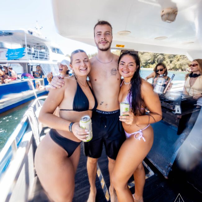 Three people in swimsuits stand closely together on a boat deck, smiling and holding canned drinks. The two women wear bikinis, while the man wears swim trunks. Other people are visible in the background on the boat, enjoying the sun and water with The Yacht Social Club Sydney Boat Hire.