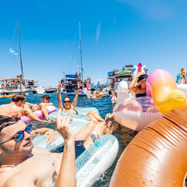 People enjoying a sunny day on the water, lounging on colorful inflatable floats. Numerous boats from The Yacht Social Club Event Boat Charters are anchored nearby, with individuals socializing and relaxing. The sky is clear and bright, adding to the festive atmosphere of the gathering.