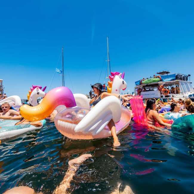 A lively water party on Sydney Harbour features people enjoying the sun on colorful inflatable floaties, including unicorns, a flamingo, and a rainbow. Boats from The Yacht Social Club can be seen in the background with others engaging in activities. An outdoor event under a clear blue sky.