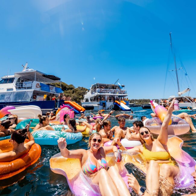 A group of people enjoying a sunny day on a lake, floating on colorful inflatable rafts. In the background, several boats from Sydney Harbour Boat Hire The Yacht Social Club and a clear blue sky. Everyone appears to be having fun, some holding drinks and smiling at the camera.