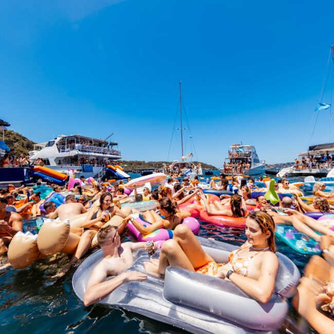 A lively crowd of people are enjoying themselves in a picturesque, sunny setting, floating on inflatable rafts and loungers in the water. Several boats from The Yacht Social Club Sydney Boat Hire are anchored nearby, creating a festive and vibrant atmosphere under a clear blue sky.