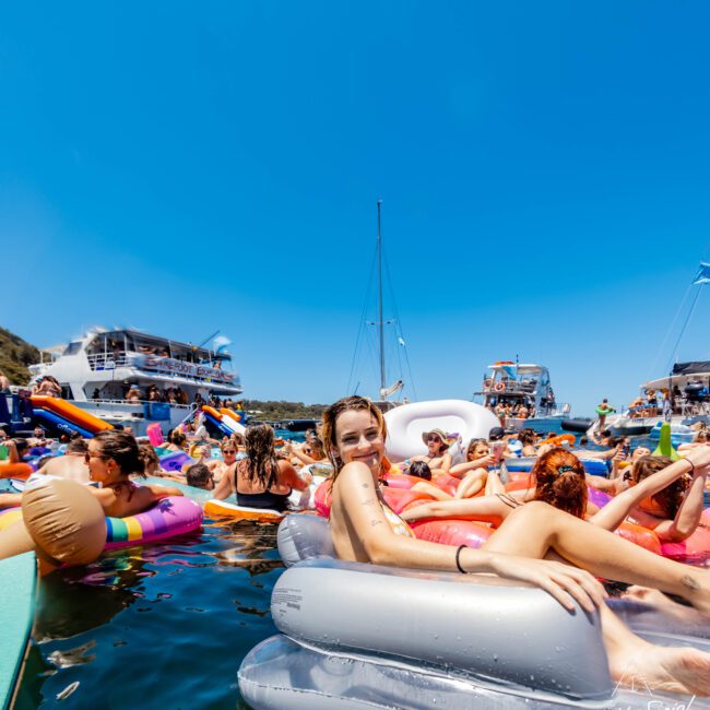 A lively scene on the water with people floating on colorful inflatable rafts and inner tubes. A young woman in the center smiles and relaxes on a gray float. Boats from Luxury Yacht Rentals Sydney add to the festive and sunny atmosphere against a clear blue sky in the background.