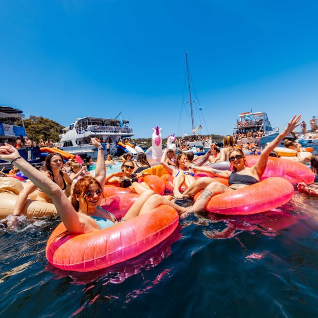 A group of people is enjoying a sunny day at Boat Parties Sydney The Yacht Social Club, floating on vibrant inflatable rafts in the water. They are smiling, raising their arms, and partying, surrounded by luxury yachts and other attendees.