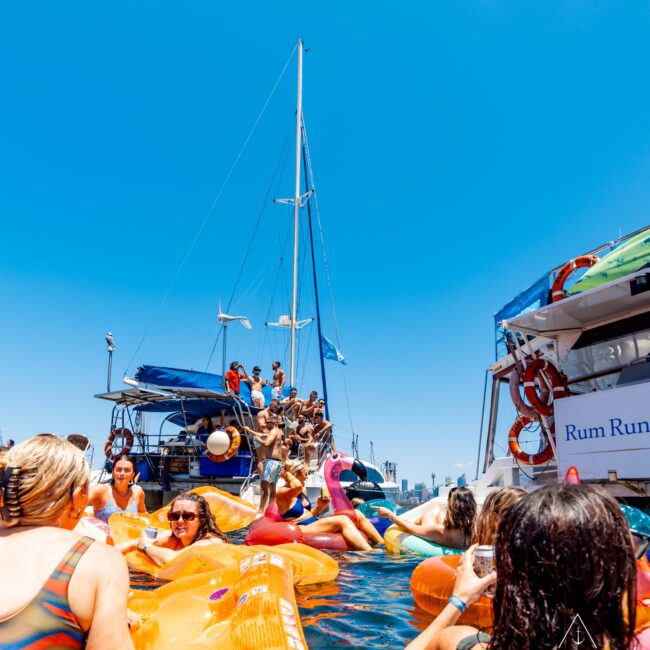 A lively scene of people enjoying a sunny day on the water, surrounded by colorful inflatable floaties. Near boats, one with a banner reading "Rum Run," some are on deck while others swim and socialize. Hosted by The Yacht Social Club Event Boat Charters, the sky is clear and blue.