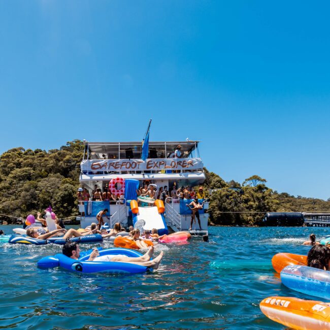 A lively scene of people enjoying a sunny day on the water with inflatable loungers and toys. Two boats, including one from The Yacht Social Club Sydney Boat Hire, are docked nearby, with passengers socializing and jumping into the water amidst lush green surroundings.