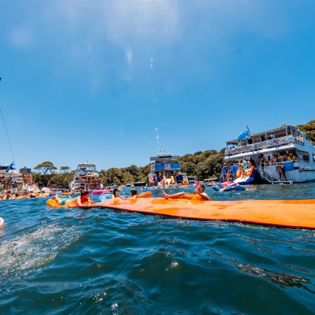 A vibrant scene of numerous people enjoying themselves on boats and floating devices in a sunny body of water. Several yachts and boats from The Yacht Social Club Event Boat Charters are anchored nearby, with people lounging on inflated mats and engaging in water activities. Trees and buildings are visible in the background.