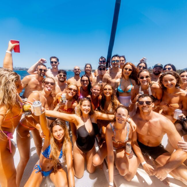 A group of young adults in swimwear are gathered on a boat, enjoying a sunny day on the water. Many are holding drinks and smiling at the camera. A bridge and blue sky are visible in the background, showcasing an event by The Yacht Social Club Sydney Boat Hire.