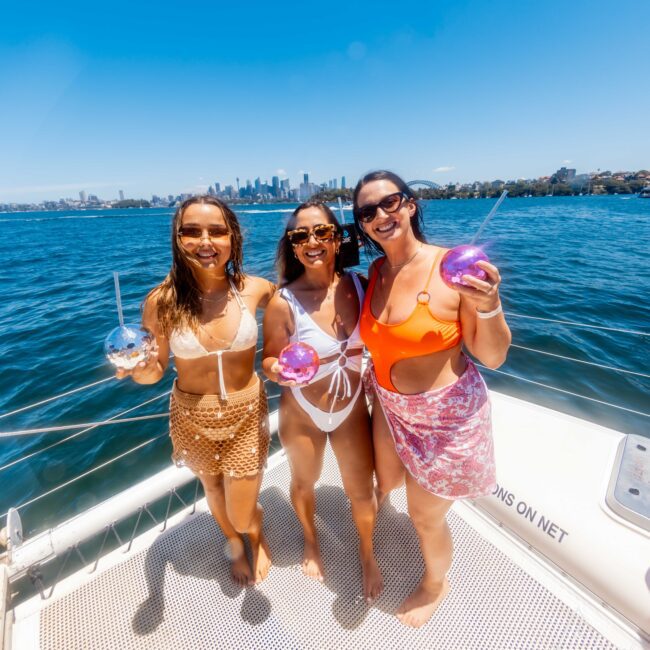 Three women in swimwear stand on a boat, smiling and holding spherical drinks. The background features a sunny, clear sky and a city skyline on the horizon with buildings and water. The women appear to be enjoying a day on the water with The Yacht Social Club Sydney Boat Hire.