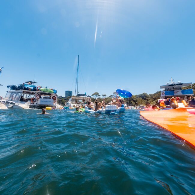 A group of people enjoying a sunny day on the water, with several boats anchored nearby and people swimming and floating on inflatable objects. In the distance, lush trees and a clear blue sky add to the vibrant summer atmosphere. Perfect for The Yacht Social Club Event Boat Charters in Sydney.