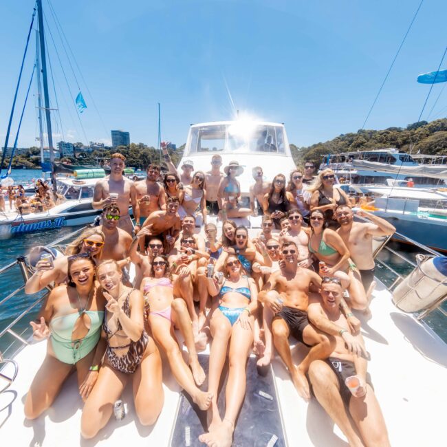 A large group of people in swimwear is gathered on the deck of a yacht, smiling and posing for a photo on a sunny day. They are enjoying a Boat Party in Sydney Harbour with The Yacht Social Club, surrounded by water, other boats, lush greenery, and a clear blue sky.