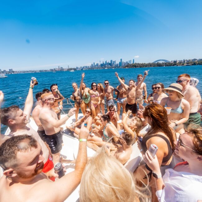 A large group of people are gathered on a boat, celebrating and raising their arms. The boat rental is part of The Yacht Social Club Sydney Boat Hire, anchored on a body of water with a cityscape, including prominent buildings and a bridge in the background. The weather is sunny and clear, creating a cheerful atmosphere.