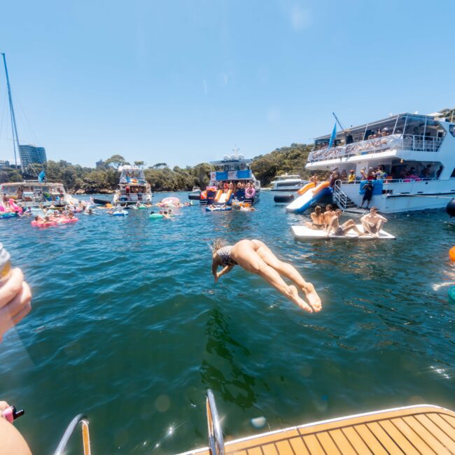 A group of people enjoy a sunny day on a lake surrounded by boats. In the foreground, a person in midair dives off a boat's deck into the water. Others relax on floaties and boats, with lush greenery in the background, creating the perfect scene for The Yacht Social Club Event Boat Charters.