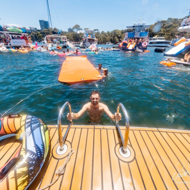 A man is climbing a ladder out of the water onto a boat deck. Several boats, inflatable rafts, and people swimming are in the background on a sunny day. The water is blue, and the atmosphere is festive and vibrant, echoing the lively spirit of The Yacht Social Club Event Boat Charters.