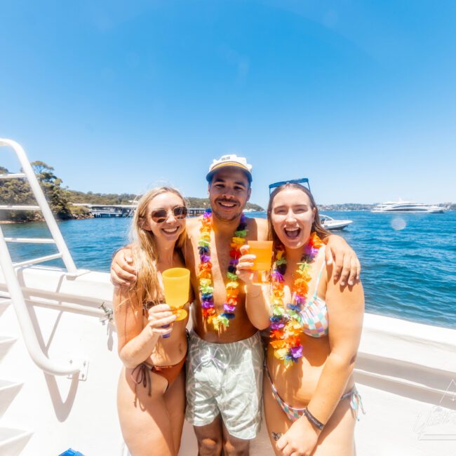 Three friends in swimsuits, smiling and holding drinks, stand together on the deck. They are all wearing colorful leis. The background features clear blue water, a distant shoreline, and a bright sunny sky – typical of Boat Parties Sydney The Yacht Social Club events.