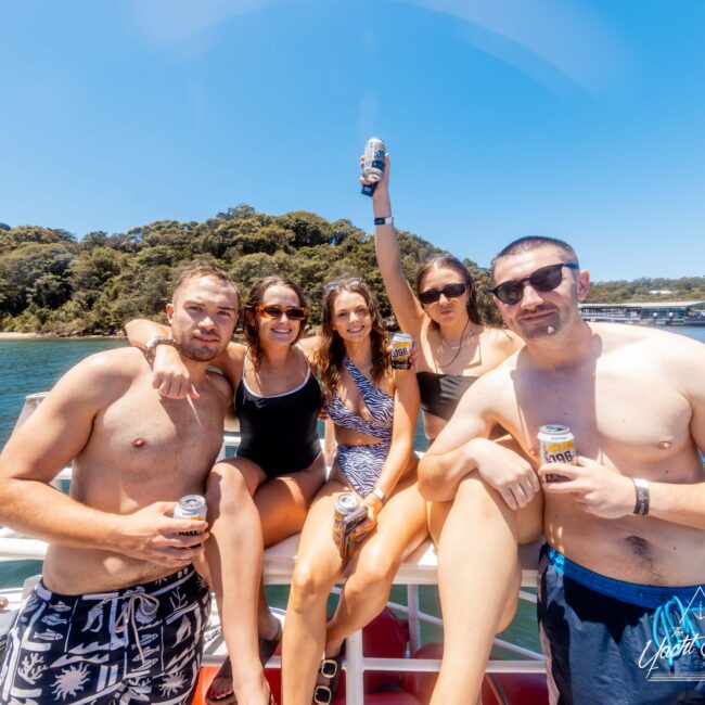 A group of five friends are gathered on a boat rental, enjoying drinks under a clear blue sky. They are in swimwear and seem to be having a fun time, with one person raising a can. There is lush greenery and calm water in the background, courtesy of Sydney Harbour Boat Hire The Yacht Social Club.