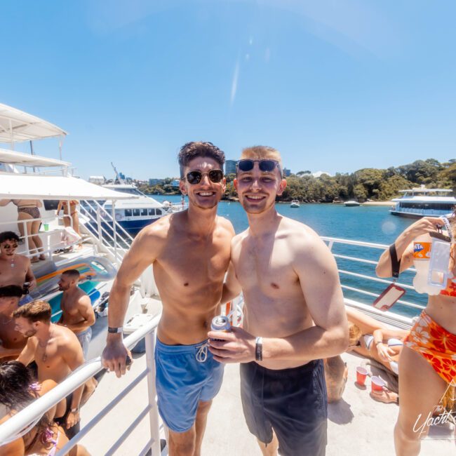 Two men smiling and posing together on a boat under the sun at The Yacht Social Club Event. One wears blue swim shorts and holds a drink, the other has on grey shorts and sunglasses. In the background, people enjoy the sunny day on the boat and in the water. Trees and another boat are visible.