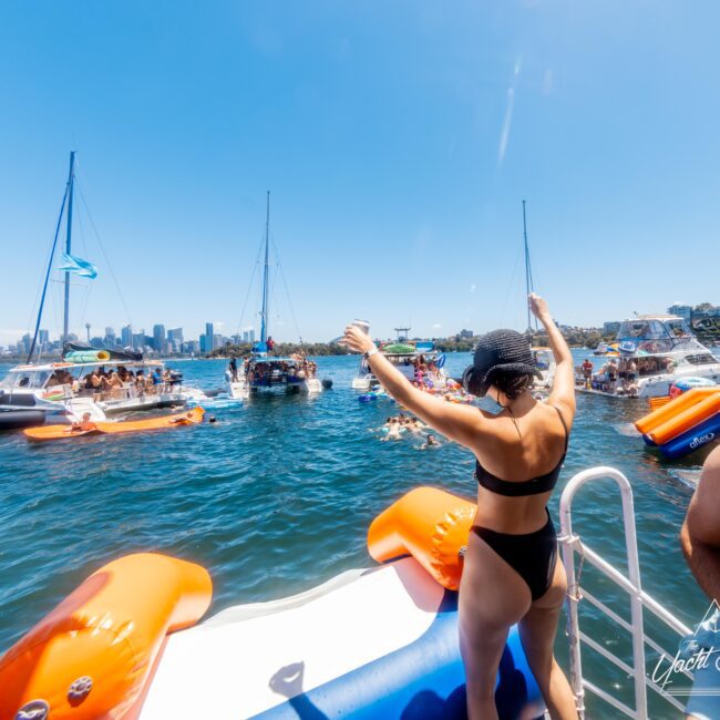 A lively scene unfolds at The Yacht Social Club, where various boats are gathered under the sun. People swim and enjoy the event, while in the foreground, a woman in a black swimsuit stands on the edge of a boat, raising her arms to embrace the festivities.