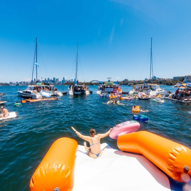 A lively scene of people enjoying a sunny day on the water with several boats anchored nearby. Some individuals are on inflatable rafts, including one person in the foreground ready to slide off an orange and white float. The city skyline is visible in the background, featuring The Yacht Social Club Sydney Boat Hire.