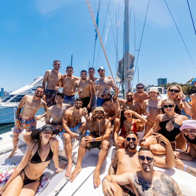 A group of 22 people, men and women, are gathered on a sailboat in their swimwear, smiling and posing for a photo. The sunny backdrop includes clear blue skies, other boats, and distant city buildings. They appear to be enjoying a daytime outing on the water with Luxury Yacht Rentals Sydney.