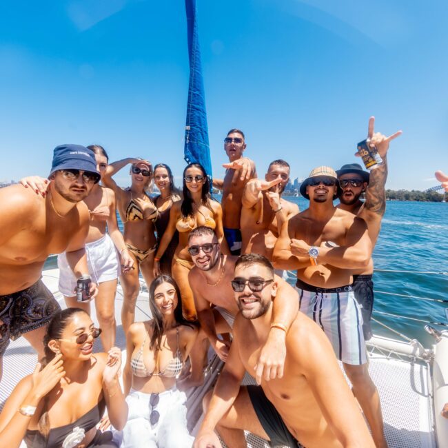 A group of people, dressed in swimwear and sunglasses, are posing and smiling on a boat during The Yacht Social Club Event Boat Charters. The sun is shining brightly, and the sea is calm in the background. Everyone appears to be enjoying their time on the water under a clear blue sky.