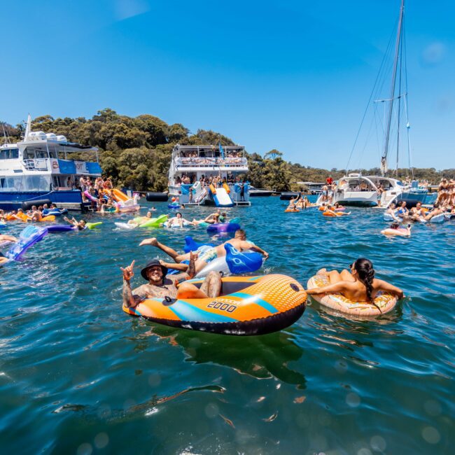 Numerous people are enjoying a sunny day on the water, floating in colorful inflatables, and swimming near anchored boats. The scene is lively with multiple boats and participants engaging in festive activities from The Yacht Social Club Event Boat Charters. The water is a deep blue under a clear sky.