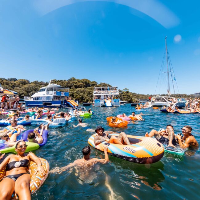 A lively scene of a yacht social club gathering in a sunny bay with Luxury Yacht Rentals Sydney. People are relaxing on colorful inflatable floats in the water, surrounded by yachts and boats. The sky is clear, and everyone is enjoying the warm weather and the festive atmosphere.