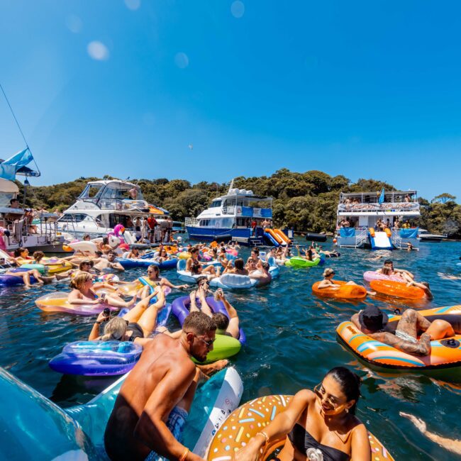 A vibrant scene of people relaxing on colorful inflatable floats in the water near several boats on a sunny day. The crowd enjoys the clear blue sky and lively atmosphere, with a forested shoreline visible in the background. This iconic setting is characteristic of Boat Parties Sydney The Yacht Social Club.