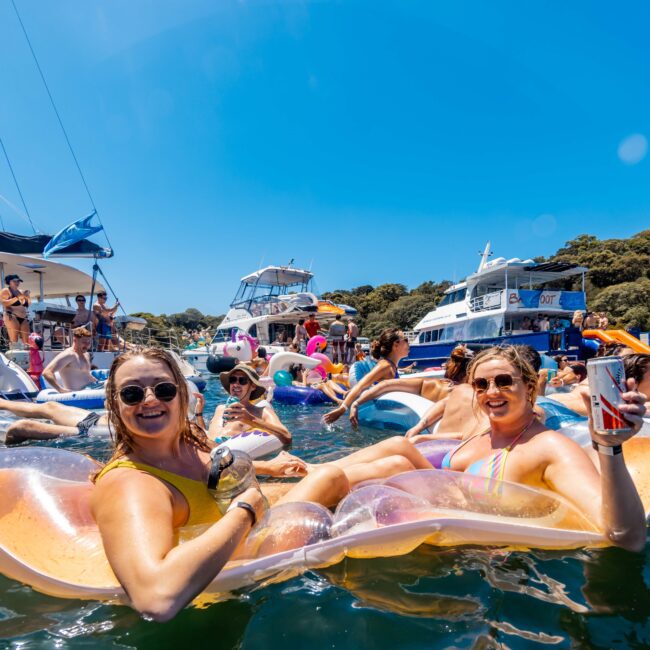 A lively scene at a boat party with people enjoying themselves in the water. Two women in swimsuits are smiling and relaxing on a colorful inflatable float. Boats and other partygoers from The Yacht Social Club Sydney Boat Hire set against a clear blue sky and vibrant greenery.