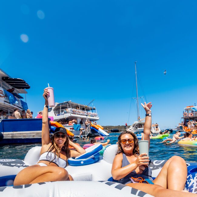 Two women relaxing on a large inflatable float in the water, each holding a drink and smiling at the camera. They are surrounded by other people on boats and inflatables, enjoying a sunny day under a clear blue sky. A seagull flies overhead, capturing the essence of Boat Parties Sydney The Yacht Social Club.