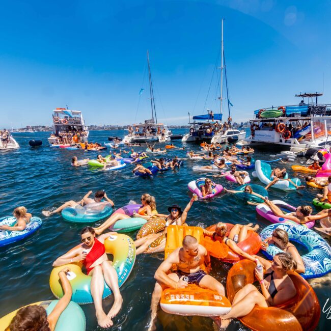 A lively gathering of people enjoying a sunny day on the water, surrounded by boats and floating on various inflatable pool floats. The clear blue sky and calm water contribute to the festive atmosphere. Hosted by The Yacht Social Club Event Boat Charters, the crowd is relaxed and having a fun time.