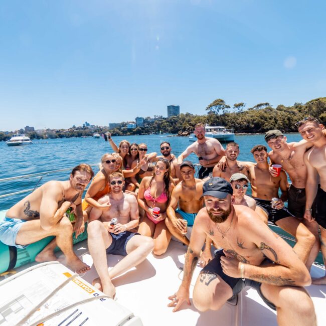 A large group of people in swimwear gather on the deck of a boat from Luxury Yacht Rentals Sydney, smiling and posing for the camera. The boat is on a body of water, with trees and buildings visible in the background. It appears to be a sunny day, and the group looks happy and relaxed.