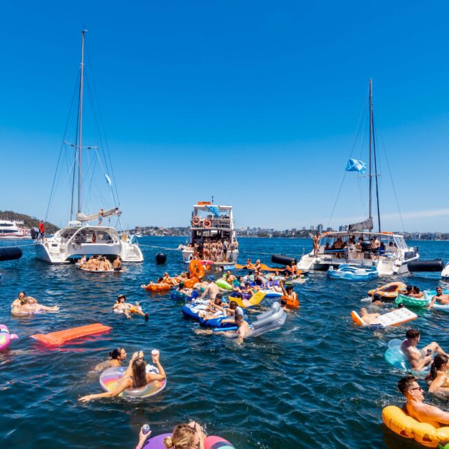 A lively scene of people enjoying a sunny day on a lake. Various colorful inflatables and boats are scattered across the water, with individuals swimming, floating, and interacting. Two sailboats and a catamaran from Sydney Harbour Boat Hire's The Yacht Social Club are anchored nearby, under a clear blue sky.