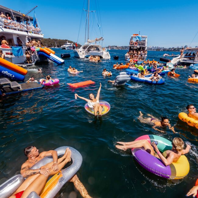 A lively scene of people enjoying a sunny day on the water, floating on inflatables in a picturesque bay. Several boats, courtesy of Luxury Yacht Rentals Sydney, are anchored nearby with more people lounging and socializing on deck. The clear blue sky enhances the festive atmosphere.