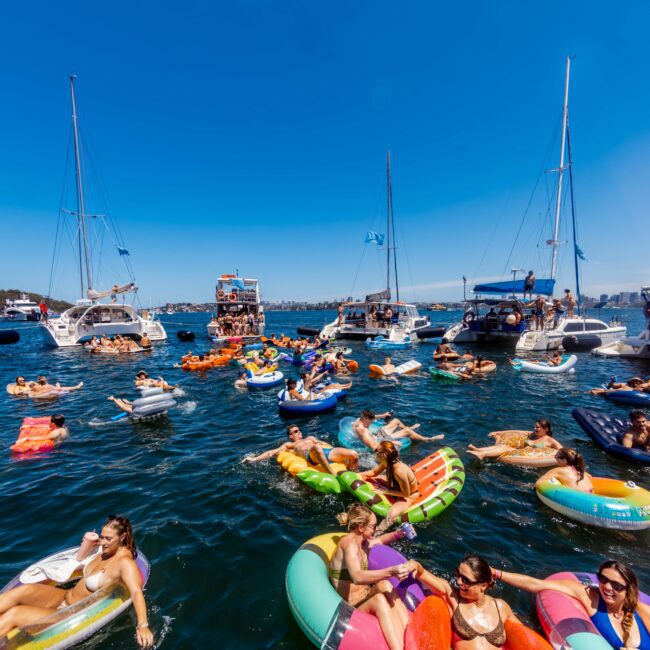 People are enjoying a sunny day on the water, floating on colorful inflatable rafts and boats. Several yachts and motorboats from The Yacht Social Club Sydney Boat Hire are anchored nearby. The water is clear and the sky is bright blue, creating a lively and festive atmosphere.