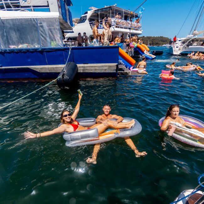 People enjoying a sunny day on the water, floating on inflatable loungers near boats. A woman in a red bikini waves at the camera, and the surrounding area is filled with others swimming and relaxing. In the background, more people are on the deck of a large yacht from Sydney Harbour Boat Hire The Yacht Social Club.