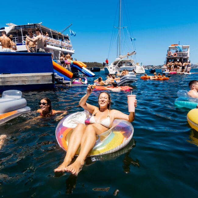 A woman in a bikini relaxes on a colorful inflatable float, holding a drink and smiling. She is surrounded by other people on floats and nearby yachts under a bright blue sky. The scene looks like a fun, sunny day on the water at The Yacht Social Club Sydney Boat Hire.