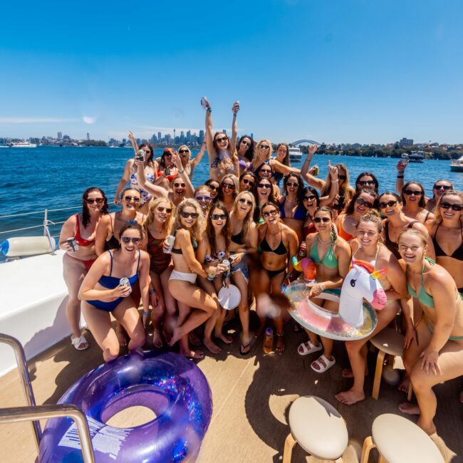 A large group of women in swimsuits are gathered on a boat under a bright blue sky. They are smiling, some with raised arms, holding drinks. The luxury yacht is anchored on a sunny day, and water and the Sydney city skyline are visible in the background. Visit Sydney Harbour Boat Hire for unforgettable experiences.